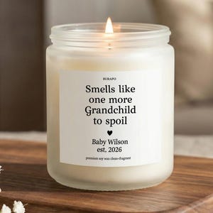 May include: A lit, frosted glass candle with the text "Smells like one more Grandchild to spoil." The candle has a white label with the name "Baby Wilson" and the year "2026." It sits on a wooden tray.