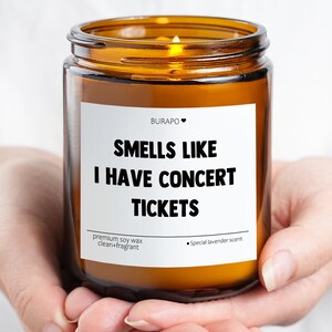 May include: A burning candle in a brown glass jar, held in cupped hands. The label says "SMELLS LIKE I HAVE CONCERT TICKETS." It is a premium soy wax candle with a special lavender scent.