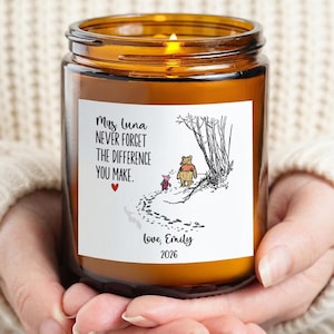 May include: A scented candle in an amber glass jar with a white label featuring a black ink illustration of Winnie the Pooh and Piglet walking. The text reads: Mrs Luna NEVER FORGET THE DIFFERENCE YOU MAKE. Love, Emily 2026, with a small red heart.