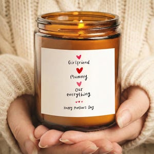 May include: A lit, amber-colored candle in a glass jar with a white label. The label reads "Girlfriend, Mummy, Our everything, Happy Mother's Day" with heart symbols. The candle is held in someone's hands.