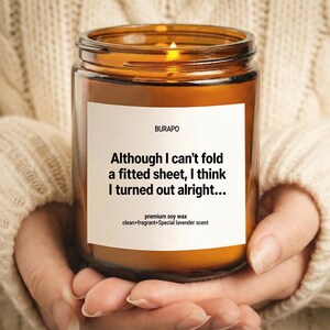 Puede incluir: Una vela de cera de soja encendida en un tarro de cristal marrón, sostenida en las manos. La vela tiene una etiqueta blanca con el texto "Although I can't fold a fitted sheet, I think I turned out alright..." y "premium soy wax".