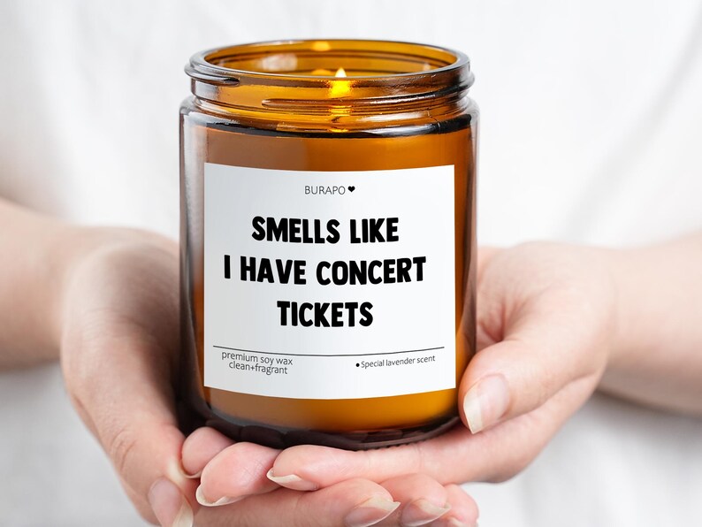 May include: A burning candle in a brown glass jar, held in cupped hands. The label says "SMELLS LIKE I HAVE CONCERT TICKETS." It is a premium soy wax candle with a special lavender scent.