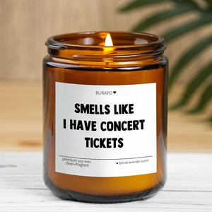 May include: An amber glass candle jar with a lit wick and a white label. The label says "Smells Like I Have Concert Tickets." The candle is made with premium soy wax and has a lavender scent.