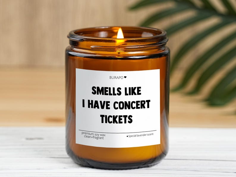 May include: An amber glass candle jar with a lit wick and a white label. The label says "Smells Like I Have Concert Tickets." The candle is made with premium soy wax and has a lavender scent.