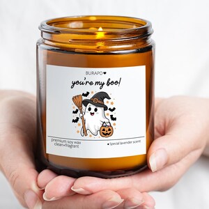 May include: A lit, amber-colored glass jar candle with a white label featuring a cartoon ghost in a witch hat holding a broom and pumpkin. The label reads "you're my boo!" and "premium soy wax".