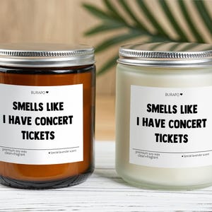 May include: Two glass jar candles with silver lids. One candle is in a brown jar, the other is in a clear jar. Each candle has a white label with the text "SMELLS LIKE I HAVE CONCERT TICKETS".