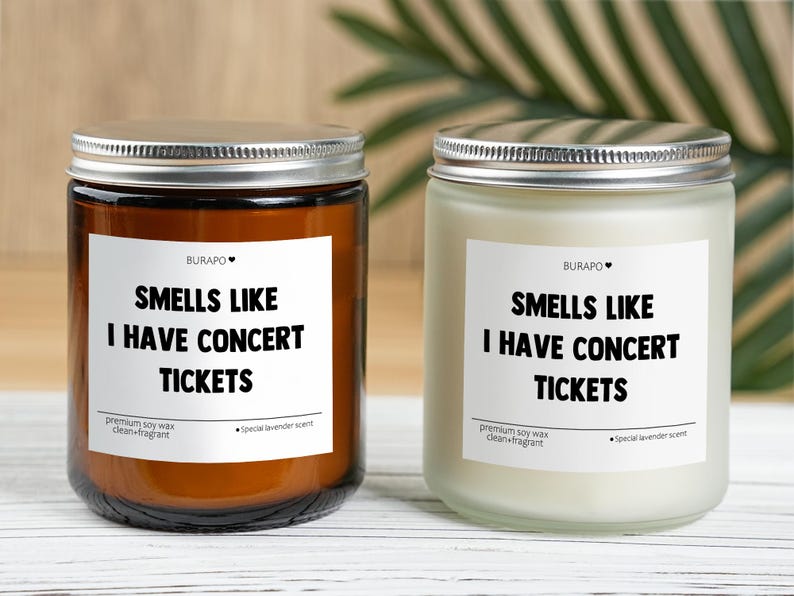 May include: Two glass jar candles with silver lids. One candle is in a brown jar, the other is in a clear jar. Each candle has a white label with the text "SMELLS LIKE I HAVE CONCERT TICKETS".