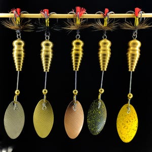 May include: Five gold fishing lures with different coloured blades. Each lure has a gold body, a silver wire, and a colourful blade. The lures are attached to a wooden dowel with red and brown accents.