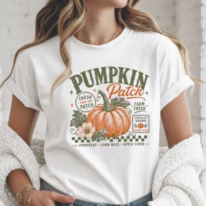 Könnte beinhalten: Ein weißes T-Shirt mit einem Grafikdruck eines orangefarbenen Kürbisses mit grünen Blättern. Der Text lautet PUMPKIN Patch, FRESH FROM THE PATCH, FARM FRESH, LOCALLY GROWN, AUTUMN PICKED, PUMPKINS, CORN MAZE und APPLE CIDER in Grün und Orange.
