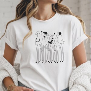 Op de afbeelding: Een wit T-shirt met korte mouwen en een zwarte lijntekening van vier windhonden die naast elkaar staan. De honden zijn afgebeeld met eenvoudige accessoires, waaronder een parelketting, een klein hoedje, een halsdoek en een bloem als haarversiering.