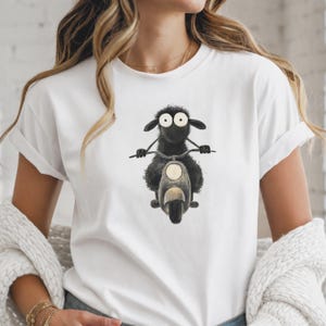 Peut inclure: Un t-shirt blanc à manches courtes orné d'une illustration graphique représentant un mouton noir pelucheux aux yeux écarquillés conduisant un petit scooter. Le mouton est placé au centre, tenant le guidon du scooter avec ses deux sabots.