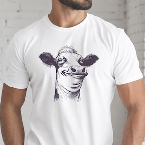 Könnte beinhalten: Ein weißes T-Shirt mit kurzen Ärmeln und einer mittigen, detaillierten schwarz-weißen Tuschezeichnung eines Kuhkopfes. Die Kuh ist mit einem ausgeprägten, breiten Lächeln dargestellt, wobei Schnauze, Ohren und Fellstrukturen sichtbar sind.