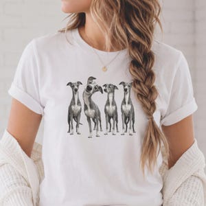 Puede incluir: Una camiseta blanca de manga corta con un estampado gráfico de cuatro galgos italianos grises y blancos de pie en fila. Los perros están representados en un estilo realista, con uno de ellos en el centro inclinando la cabeza hacia un lado.