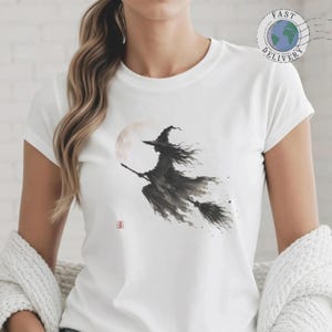Può includere: Una t-shirt bianca a maniche corte con un'illustrazione a inchiostro nero di una strega che vola su una scopa davanti a una luna pallida. La grafica include un piccolo timbro quadrato rosso vicino alla parte inferiore sinistra della strega.