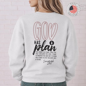 Könnte beinhalten: Ein hellgraues Langarm-Sweatshirt mit dem Schriftzug GOD HAS A PLAN in großen rosa und schwarzen Buchstaben auf der Rückseite. Darunter befindet sich der Bibelvers Jeremiah 29:11 in schwarzem Druck, der von Plänen für Hoffnung spricht.