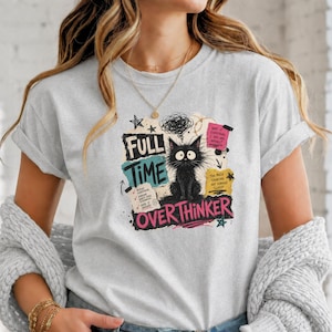 Op de afbeelding: Lichtgrijs t-shirt met een zwarte kat illustratie en de tekst "FULL TIME OVERTHINKER". Het ontwerp bevat handgeschreven notities en krabbels, wat een grillige uitstraling geeft.