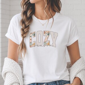 Könnte beinhalten: Ein weißes kurzärmeliges T-Shirt mit dem aufgedruckten Text COZY season auf der Vorderseite. Die Buchstaben von COZY sind mit einem beigefarbenen und weißen Karomuster sowie Blumenillustrationen in Orange, Grün und Weiß gefüllt.
