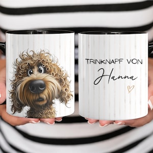 Könnte beinhalten: Ein weißer Keramikbecher mit schwarzem Henkel und schwarzem Innenraum, der auf einer Seite eine Illustration eines braunen Hundes mit lockigem Fell zeigt und auf der anderen Seite den Text "TRINKNAPF VON Hanna" mit einem kleinen Herzsymbol.
