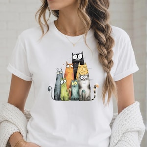 Könnte beinhalten: Ein weißes kurzärmeliges T-Shirt mit einer grafischen Illustration von sieben Cartoon-Katzen in verschiedenen Farben und Größen, darunter Schwarz, Orange, Gelb, Grün und Grau, alle mit großen Augen und markanten Gesichtsausdrücken.