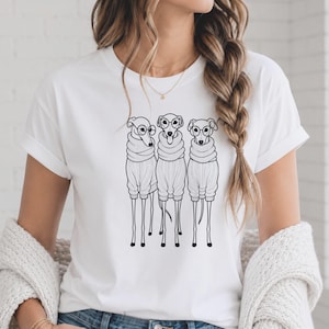 Op de afbeelding: Een wit t-shirt met korte mouwen en een zwarte lijntekening van drie windhonden die naast elkaar staan. Elke hond draagt een geribbelde coltrui en een ronde bril, waarbij de middelste hond zijn tong uit de bek heeft hangen.