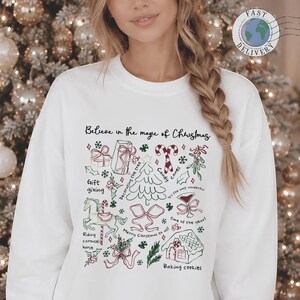 Może przedstawiać: Biała bluza z długim rękawem z napisem Believe in the magic of Christmas oraz różnymi świątecznymi ilustracjami, takimi jak koń na karuzeli, laski cukrowe, domek z piernika, prezenty i choinka z czerwonymi i zielonymi akcentami.