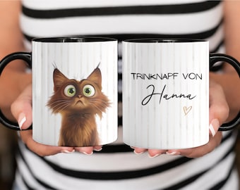 May include: A white ceramic mug with a black handle and interior, featuring a cartoon brown cat with large green eyes on one side and the text "TRINKNAPF VON Hanna" with a small heart symbol printed on the other side against a striped background.