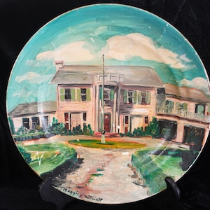 May include: A decorative plate featuring a hand-painted landscape of a house with a light pink facade, green shutters, and a central flagpole. The sky is painted in shades of blue and white. The plate is displayed on a black stand.