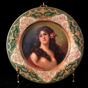 May include: A decorative round plate featuring a portrait of a woman with long brown hair and a rose. The plate has a green border with gold accents and floral patterns, and is displayed on a gold stand.