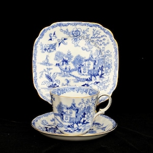 Może przedstawiać: Zestaw do herbaty z porcelany w stylu vintage z niebiesko-białym wzorem Willow. Zestaw zawiera filiżankę, spodek i kwadratowy talerz. Krawędzie filiżanki, spodka i talerza są wykończone złotem.