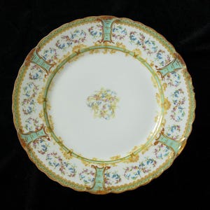 May include: A white porcelain plate with a scalloped edge, featuring a floral design in blue, yellow, and pink. The plate has a light green border with gold accents and a central floral bouquet.