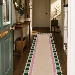 Op de afbeelding: Een rechthoekig gangtapijt met een crèmekleurig midden en een geometrische rand van roze en groene vormen. Het tapijt ligt op een donkere houten vloer in een goed verlichte gang, met uitzicht op een deur en een spiegel.