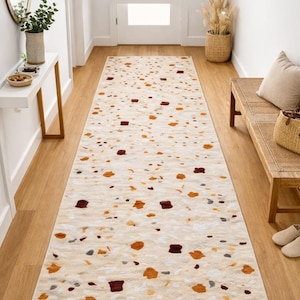 Könnte beinhalten: Ein rechteckiger Läuferteppich mit cremefarbenem Hintergrund und einem Terrazzo-Muster aus braunen, orangefarbenen und grauen Formen. Der Teppich liegt in einem Flur mit Holzböden und einer Bank.
