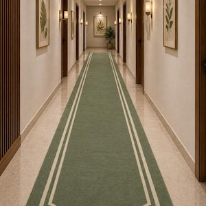 May include: A long, rectangular green rug with a white border and trim, placed in a hallway. The rug is centered in the hallway, which has multiple closed brown doors, framed botanical prints, and wall sconces.