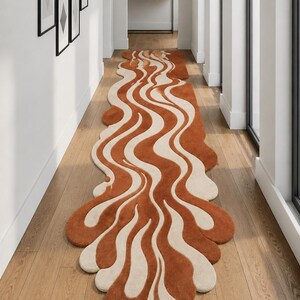 May include: A long, abstract rug with a wavy design in shades of rust and cream. The rug is placed in a hallway with hardwood flooring and white walls. The rug's unique shape and color palette create a modern, artistic aesthetic.