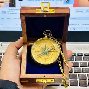 Könnte beinhalten: Ein antiker Messingkompass mit goldener Kette, untergebracht in einer Holzbox mit blauem Samtinterieur. Das Kompasszifferblatt zeigt Richtungsmarkierungen und das Wort "LORD". Die Box ist geöffnet und zeigt den Kompass.