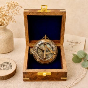 May include: A brass sundial compass with a folding gnomon and engraved markings, housed in a square wooden box with a dark blue velvet interior lining and a gold-toned metal latch. The box lid is open, revealing the compass inside.