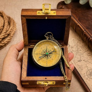 Op de afbeelding: Een vintage messing kompas met ketting, gepresenteerd in een houten kistje met een messing sluiting. De kompas wijzerplaat heeft richtingsmarkeringen en een groene edelsteen. De kist is gevoerd met blauw fluweel.