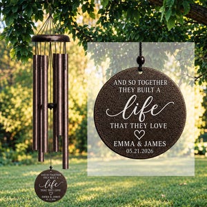 Peut inclure: Un carillon à vent de couleur bronze composé de cinq tubes cylindriques et d'un pendentif circulaire. Le pendentif porte l'inscription gravée : AND SO TOGETHER THEY BUILT A life THAT THEY LOVE, un cœur, EMMA & JAMES, et 05.21.2026.