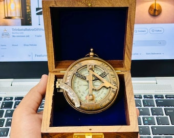 Könnte beinhalten: Ein antiker nautischer Kompass aus Messing, eingebettet in einer Holzkiste mit blauem Samtinnenraum. Der Kompass weist aufwendige Details und einen Klappdeckel mit Messingverschluss auf. Die Kiste ist aus dunklem Holz mit poliertem Finish.