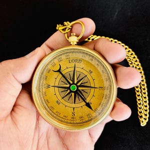 Puede incluir: Una brújula de latón vintage con una cadena dorada. La esfera de la brújula presenta marcas direccionales, una aguja y la palabra "LORD". La brújula se sostiene en una mano sobre un fondo oscuro.