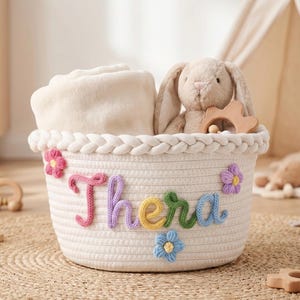 Puede incluir: Una cesta blanca tejida con el nombre "Thera" en letras de hilo colorido, decorada con flores. La cesta contiene una manta color crema, un conejito de peluche y un juguete de madera. Tema de guardería.