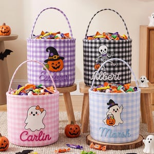 May include: Four cylindrical fabric Halloween candy buckets with gingham patterns in purple, black, pink, and light blue. Each bucket features a handle, a unique embroidered name, and a festive graphic including a pumpkin, skeleton, or ghost.