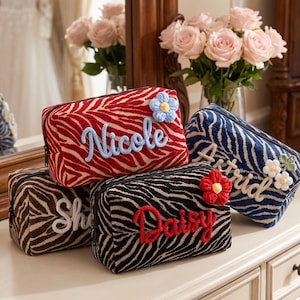 May include: Four rectangular zippered pouches with a zebra stripe pattern in red, blue, brown, and black. Each pouch features a personalized name in cursive script and a small crocheted flower appliqué attached to the front surface of the fabric.