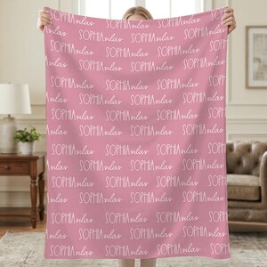 Può includere: Una coperta rosa con un motivo ripetuto del nome "SOPHIA silas" in scrittura bianca. La coperta è tenuta in alto, mostrando il design. La coperta è fatta di un materiale morbido.