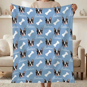 May include: A light blue blanket featuring a repeating pattern of black and white Boston Terrier dog faces, white dog bones, and the word "Satan" in a script font. The blanket is held up by a person.