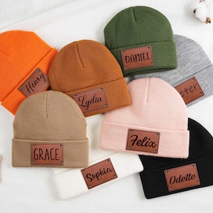 May include: A selection of colourful beanies, including orange, brown, green, grey, pink, beige, black, and white. Each hat has a brown leather patch with a name. Names include Henry, Lydia, Daniel, Peter, Felix, Grace, Sophia, and Odette.