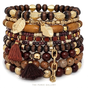 May include: A stack of multi-strand bracelets featuring dark brown wooden beads, smooth tan wooden cylinders, metallic gold-tone beads, and small red square beads. The set includes two gold-tone leaf charms, two dark red tassels, and a gold-tone chain link.