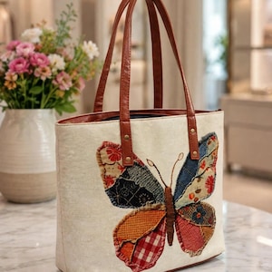 Puede incluir: Un bolso tote de color crema con una gran aplicación de mariposa de múltiples texturas en la parte delantera. La mariposa está compuesta por varios parches de tela con patrones en rojo, naranja, azul y negro. El bolso tiene dos asas de piel sintética marrón.