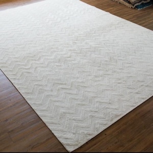 May include: A large, rectangular off-white rug with a repeating chevron pattern. The rug is made of a soft, textured material and is placed on a dark brown hardwood floor. The rug's design adds a subtle, yet stylish, element to the room.
