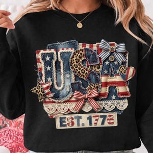 Puede incluir: Sudadera negra con un diseño patriótico "USA" que combina mezclilla, estampado de leopardo y patrones de la bandera estadounidense. El diseño incluye estrellas, lazos y el texto "EST. 1776". Con ribete de encaje.